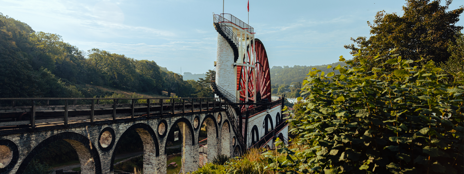 The Great Laxey Wheel on the Isle of Man. The world's largest surviving waterwheel of its kind. An arched aqueduct leads towards spiral steps next to the giant red water wheel, situated in a green valley.